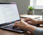 Close-up of hands typing on a laptop at a clean desk in soft natural light, with the screen content blurred and a closed notebook, small plant, and mug in the background.